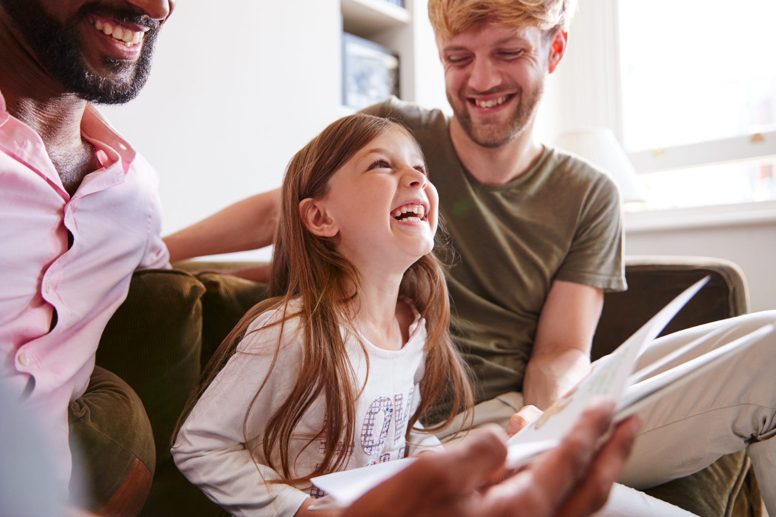 A gay male couple sit with their young daughter, laughing and reading a story