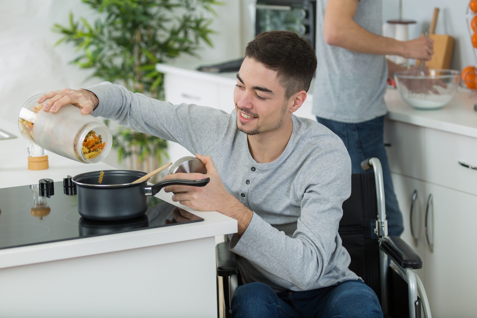A man in a wheelchair cooking at the stove in a kitchen, tipping pasta into a sauce pan