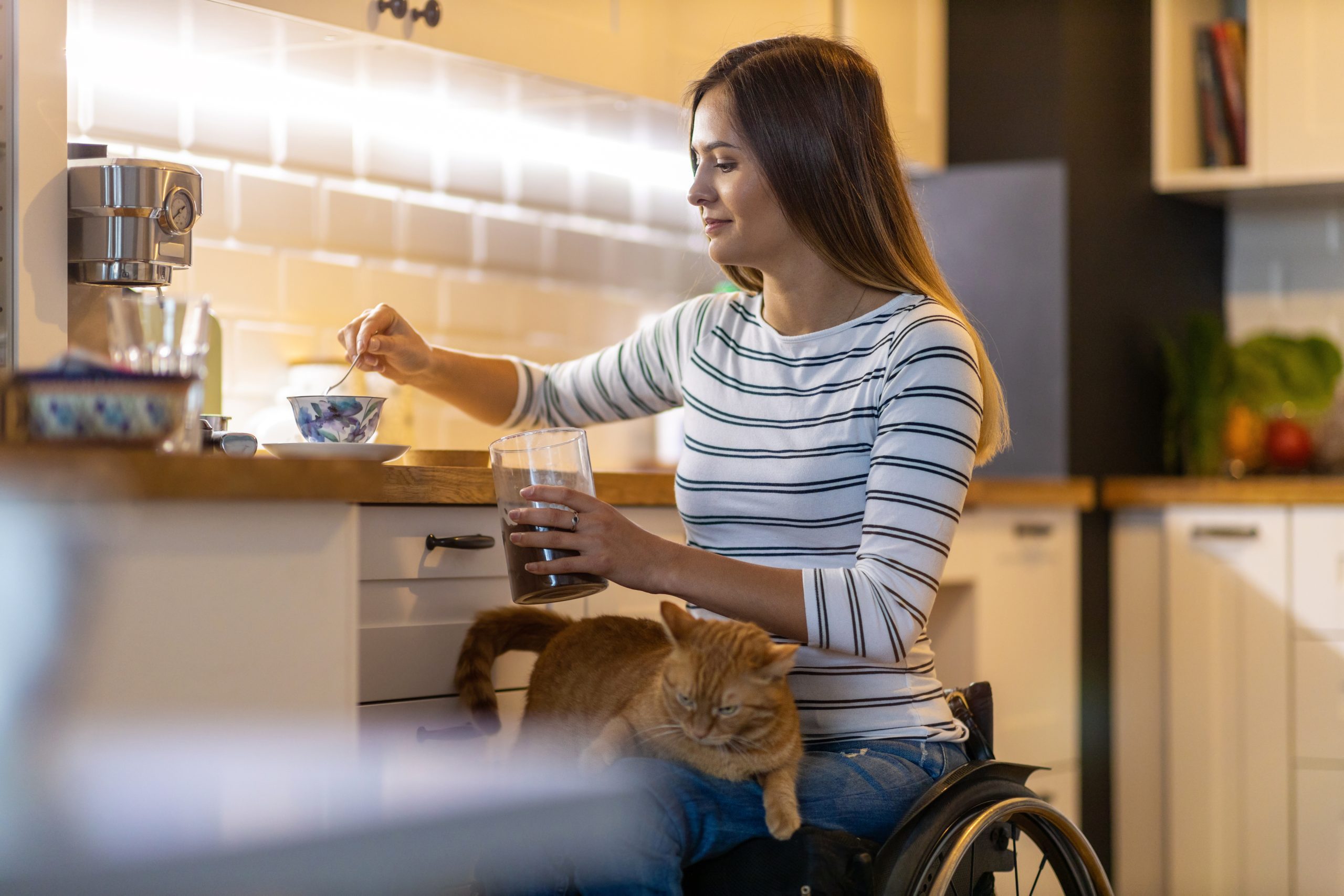 Woman in wheelchair making tea in the kitchen with her cat on her lap